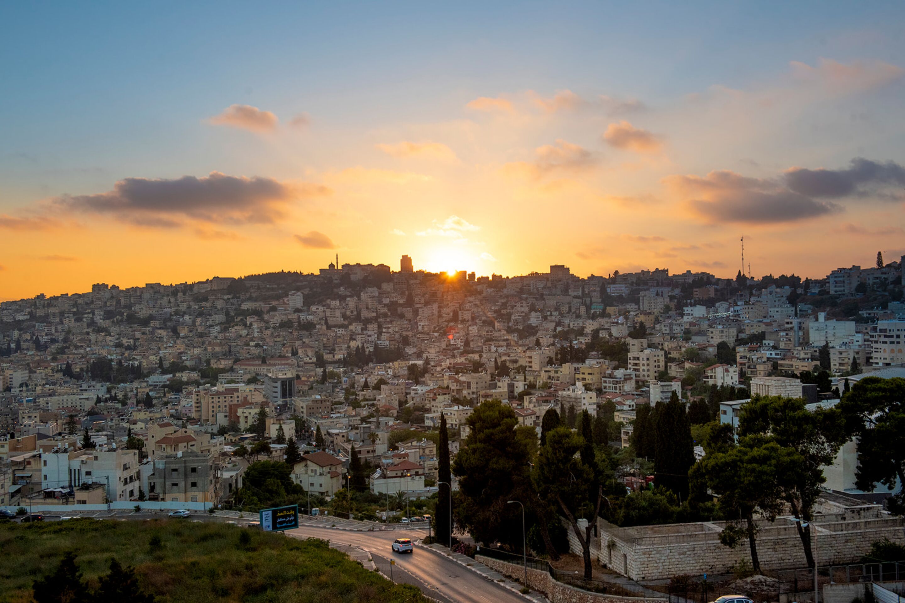 A cityscape at sunset with a dense urban area, a road with vehicles, and a clear sky.