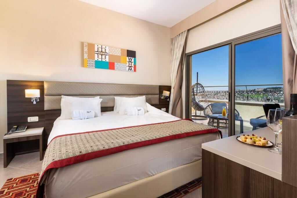 A hotel room with a large bed, a desk with a plate of food and a glass, and a balcony with a view of the city.
