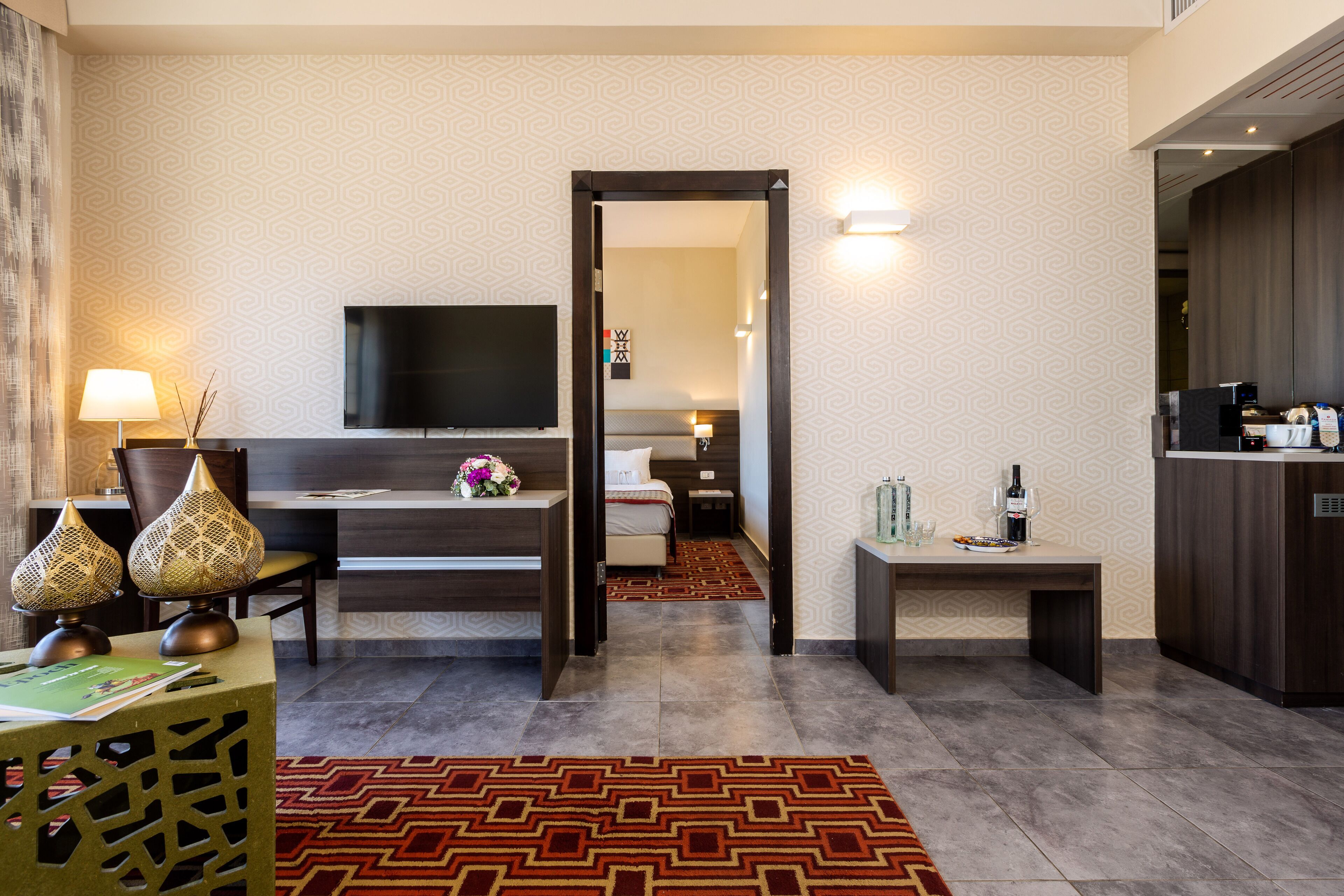 Een moderne hotelkamer met een flatscreen-tv, een bureau met een lamp, een bed en een klein tafeltje met een fles.