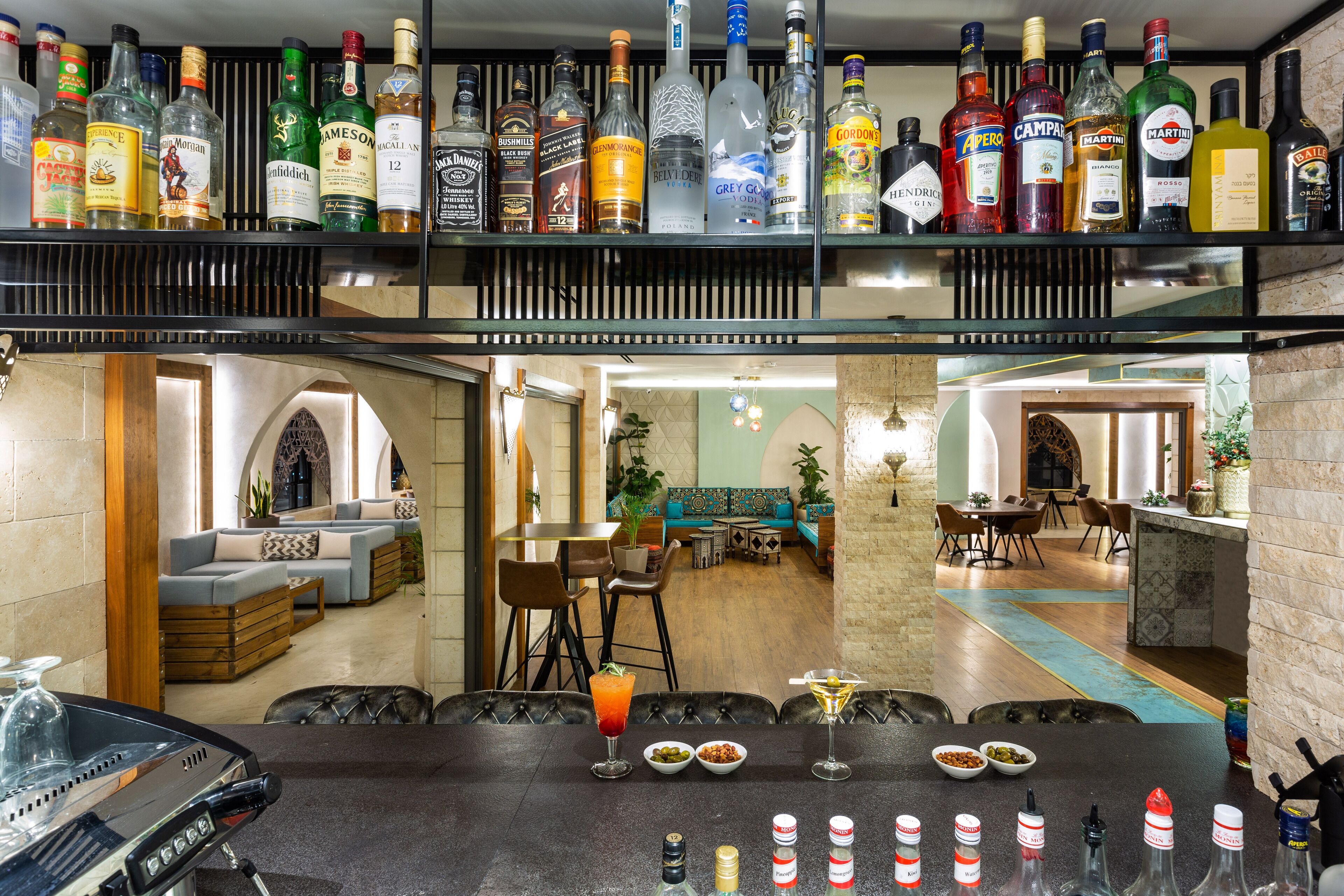 Una zona de bar con una variedad de botellas de licor en estantes, una barra con vasos y condimentos, y un área de descanso con sofás y mesas.