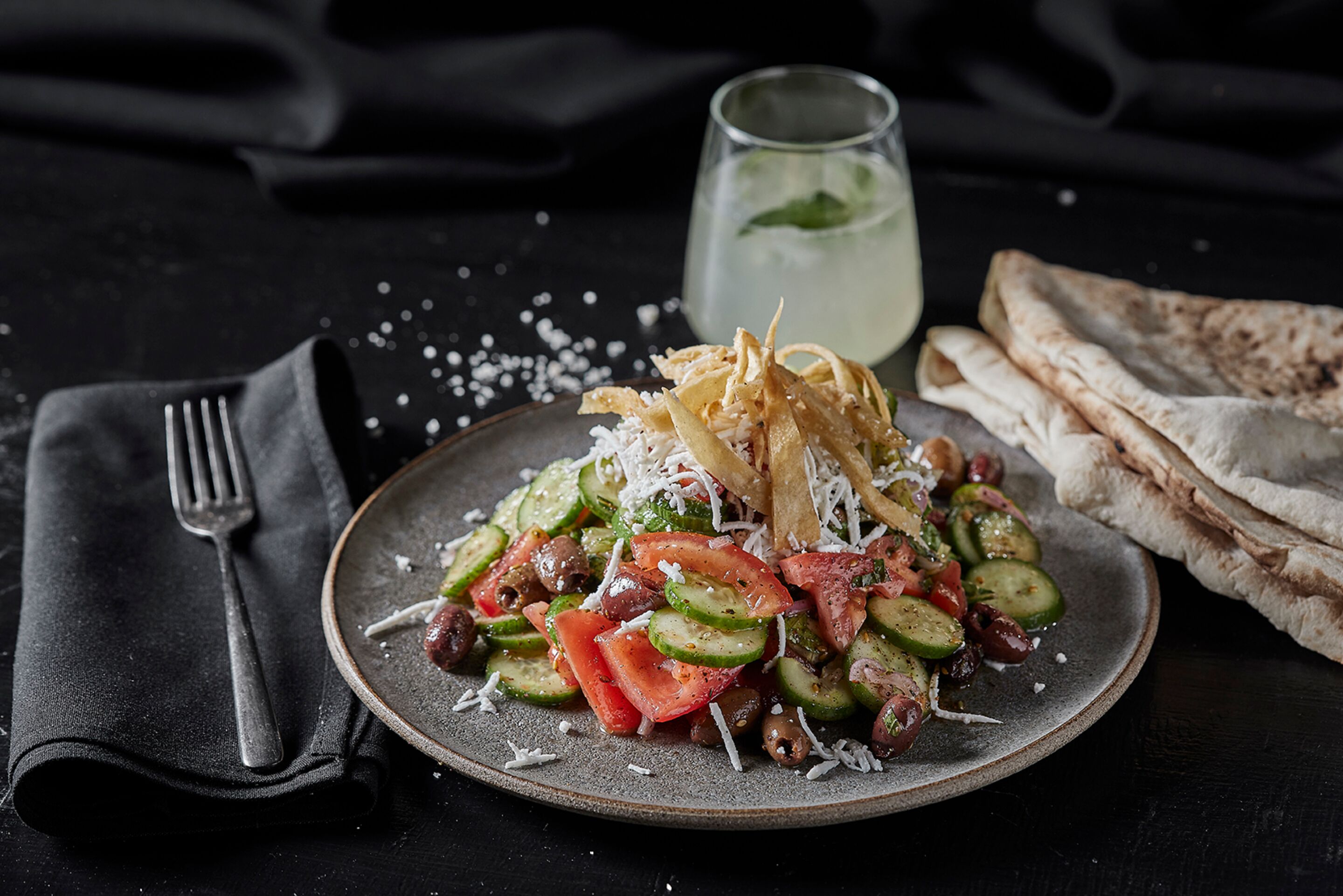 A plate of vegetables with cheese and olives, a glass of water, and flatbreads on a dark surface.