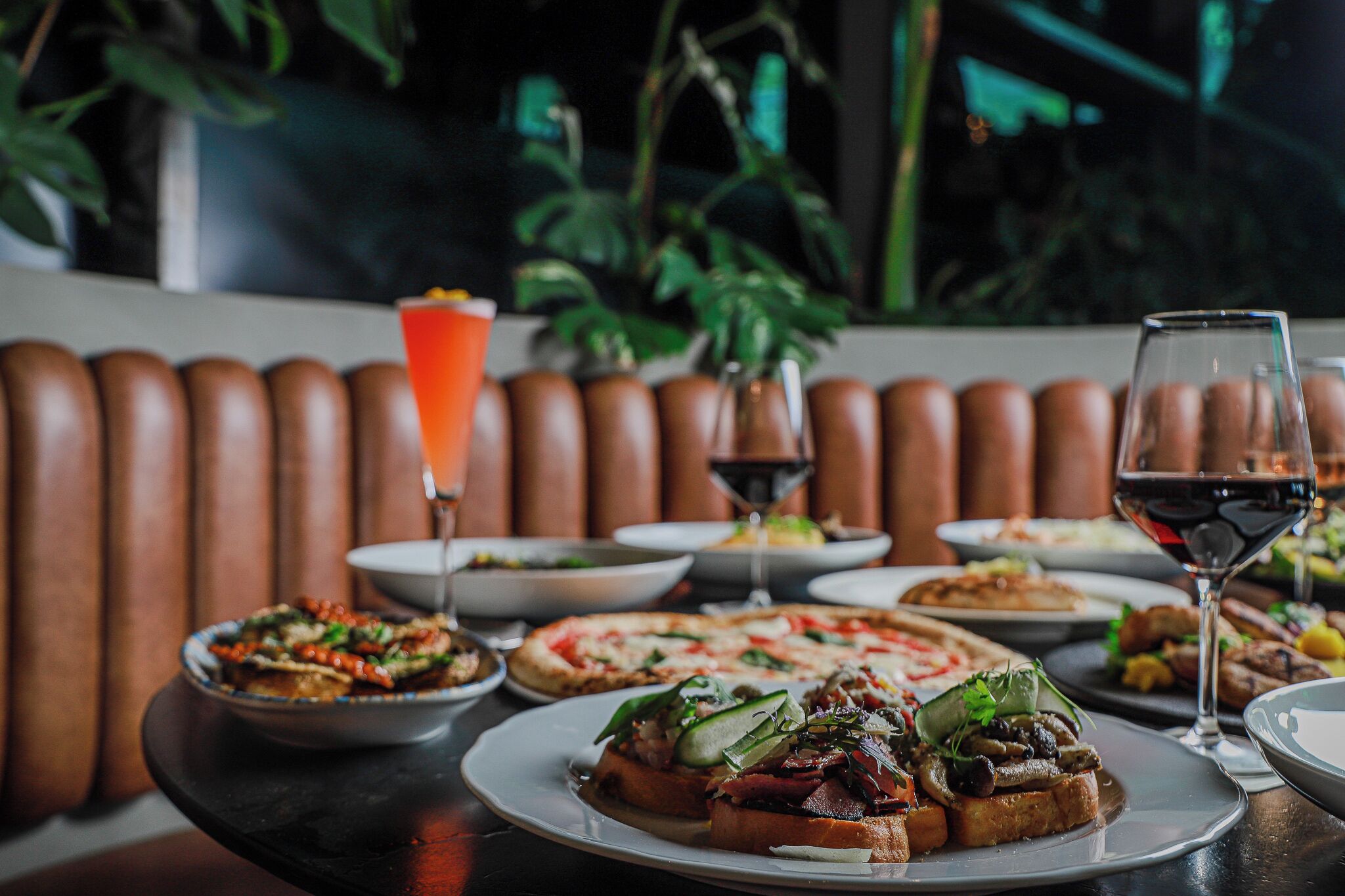 A table set with various dishes, including pizzas and salads, and two glasses of red wine.