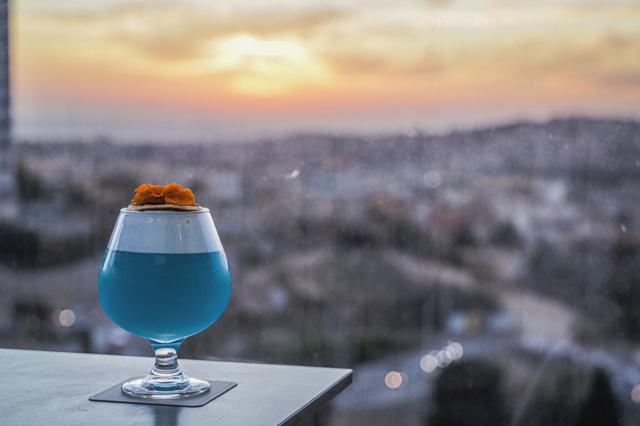 Een cocktailglas met blauwe vloeistof en een oranje garnering op een onderzetter, voor een onscherpe stadsgezicht en zonsondergang op de achtergrond.