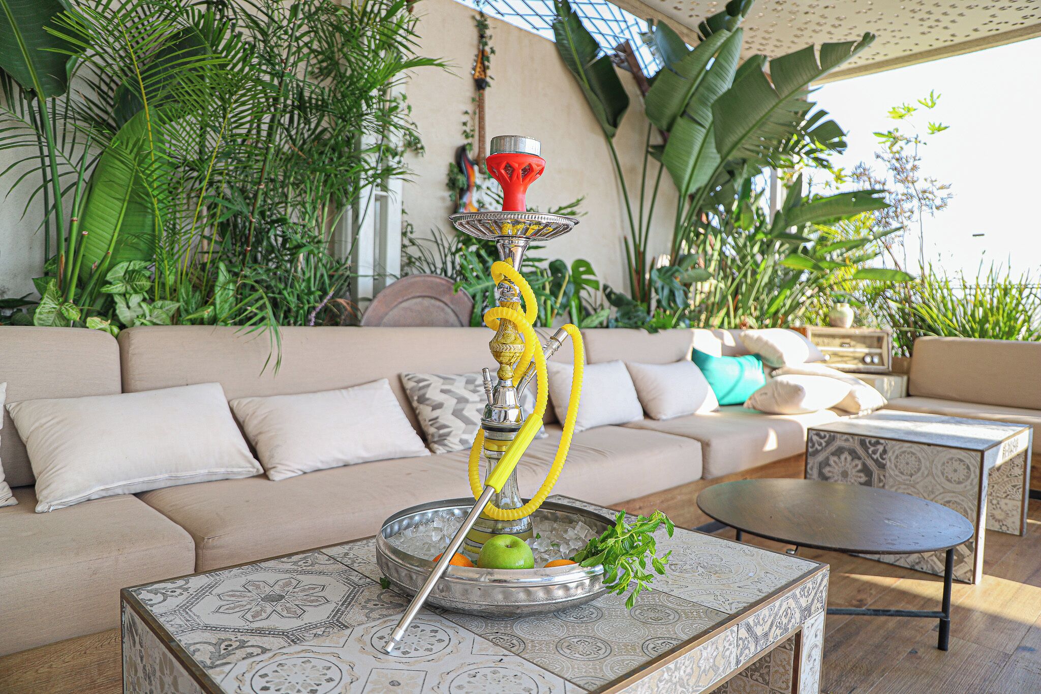 A hookah on a patterned table with a sofa and decorative plants in the background.