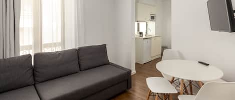 A modern living room with a grey sofa, a dining table with chairs, a kitchen in the background, and a flat-screen TV mounted on the wall.