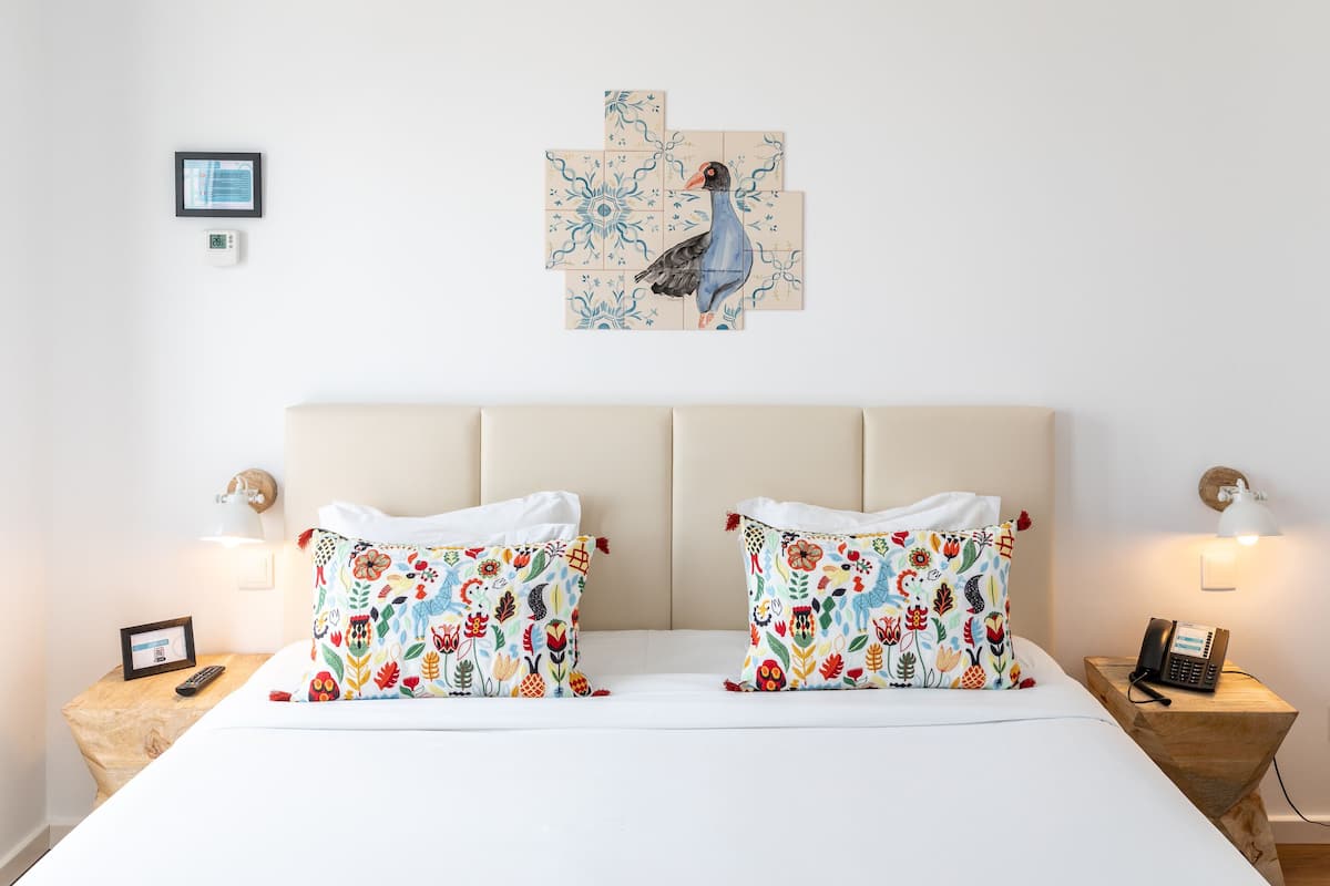 Une chambre d’hôtel avec un lit, deux oreillers à motifs floraux, une œuvre d’art représentant un oiseau accrochée au mur, des lampes de chevet, un téléphone sur la table de chevet et un petit tableau encadré posé sur la même table.