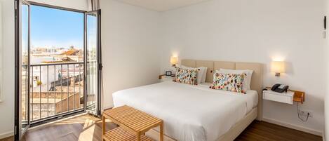 A modern hotel room with a large bed, a wooden bench, a bedside table with a phone, and a view of the city through a large window.
