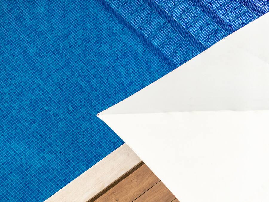 Una terraza de madera con piscina, una silla de descanso y una toalla azul, junto a una pared blanca.