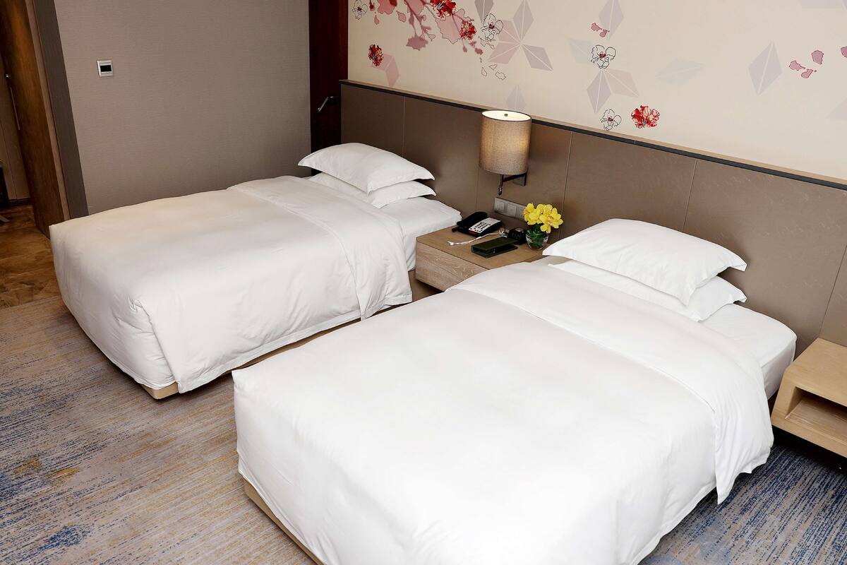 A hotel room with two single beds, a nightstand with a lamp, a phone, and a vase with flowers.