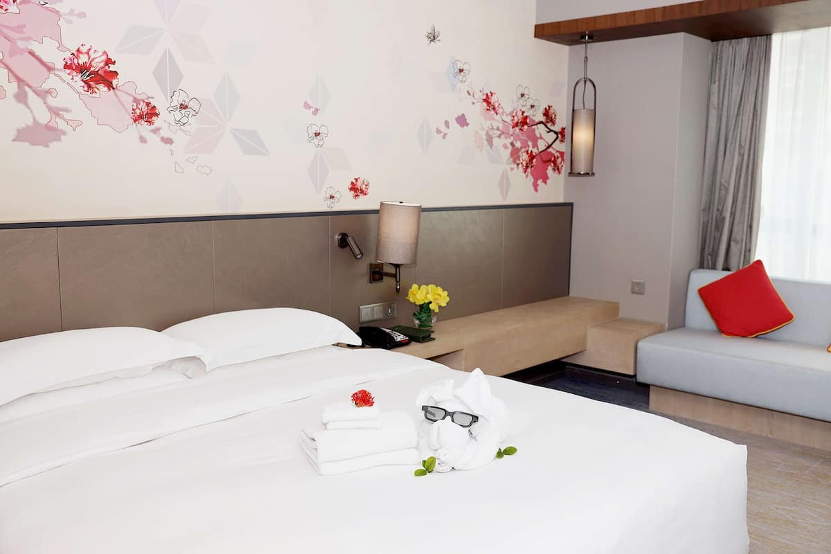 A hotel room with a neatly made bed, a sofa with a red pillow, and a wall with floral and geometric patterns.
