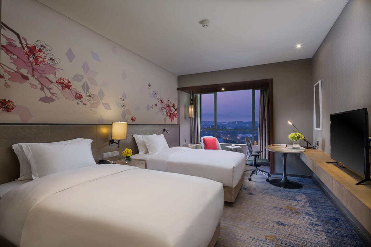 A hotel room with two beds, a TV, a desk with a chair, a small table with a vase of flowers, and a large window offering a city view.