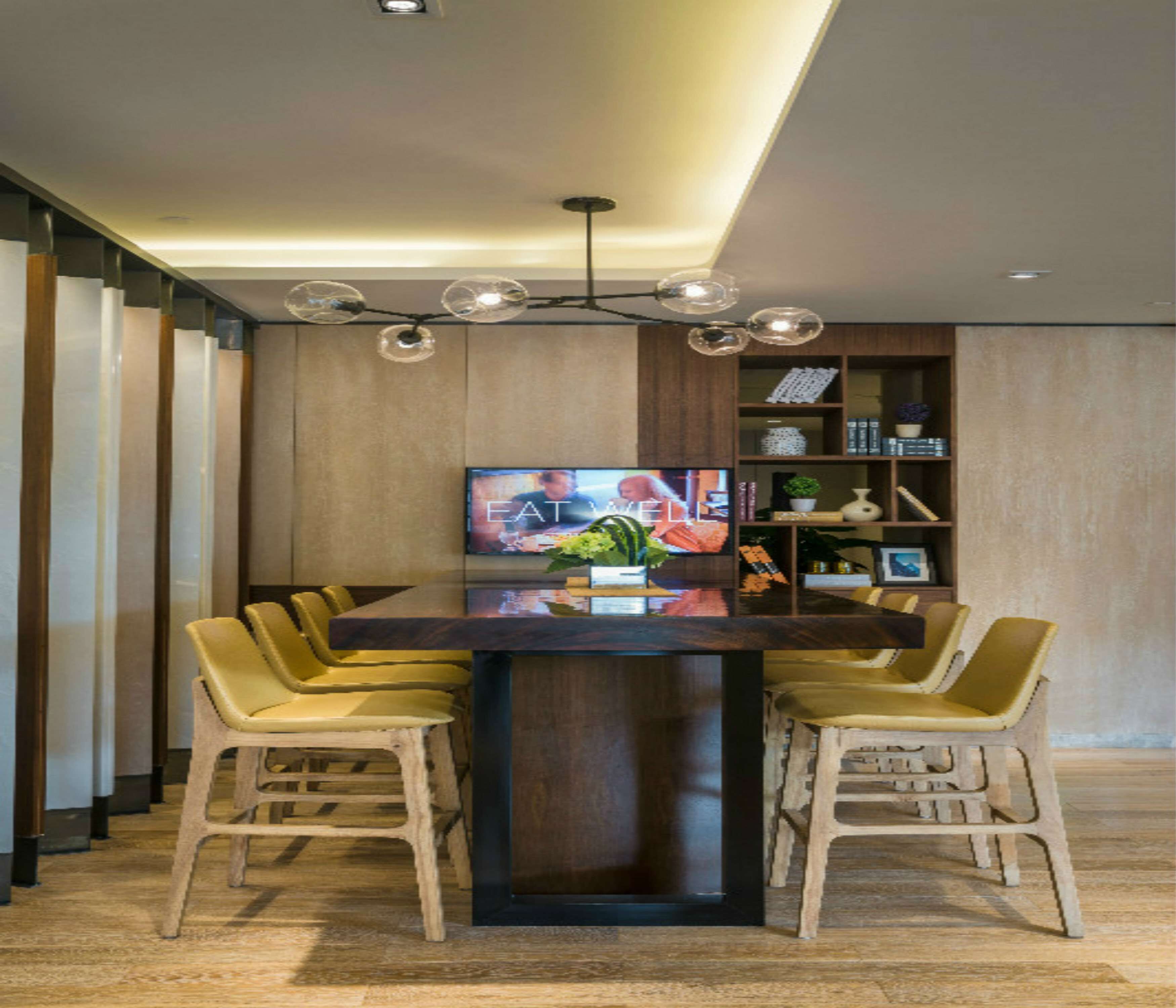 A modern dining area with a large wooden table, six yellow chairs, a television displaying a cooking show, and a ceiling light fixture.