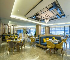 A modern hotel lobby with a high ceiling, large windows, and a mix of seating areas including a blue sofa and yellow chairs.