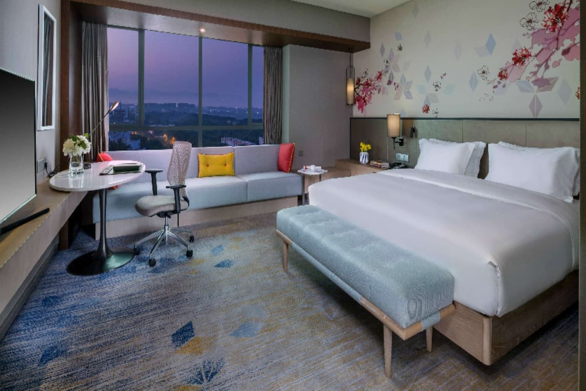 A hotel room with a large bed, a sofa, a desk with a chair, a nightstand, and a view of the city at dusk.