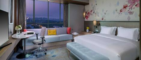 Habitación de hotel con una cama grande, un sofá, un escritorio con una silla, una mesita de noche y vistas a la ciudad al anochecer.