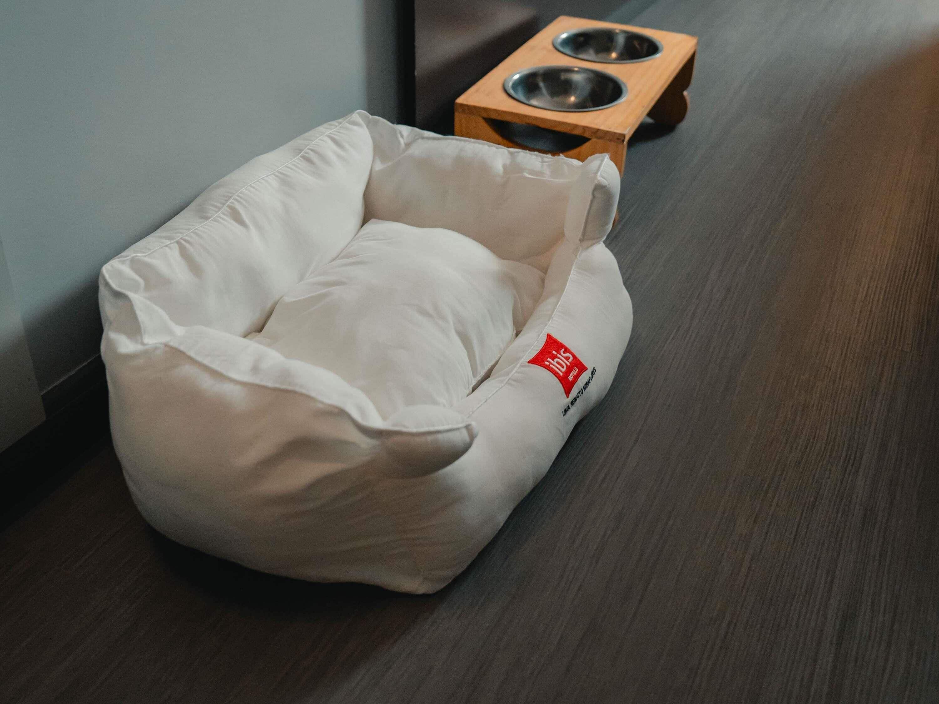A white pet bed with a red logo, placed on a wooden floor next to a pet feeding station.