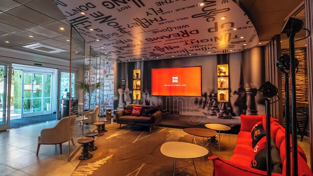 Een moderne lobby met een gebogen plafond, voorzien van inspirerende citaten, een groot scherm waarop een logo staat afgebeeld, en een zithoek met banken en tafels.