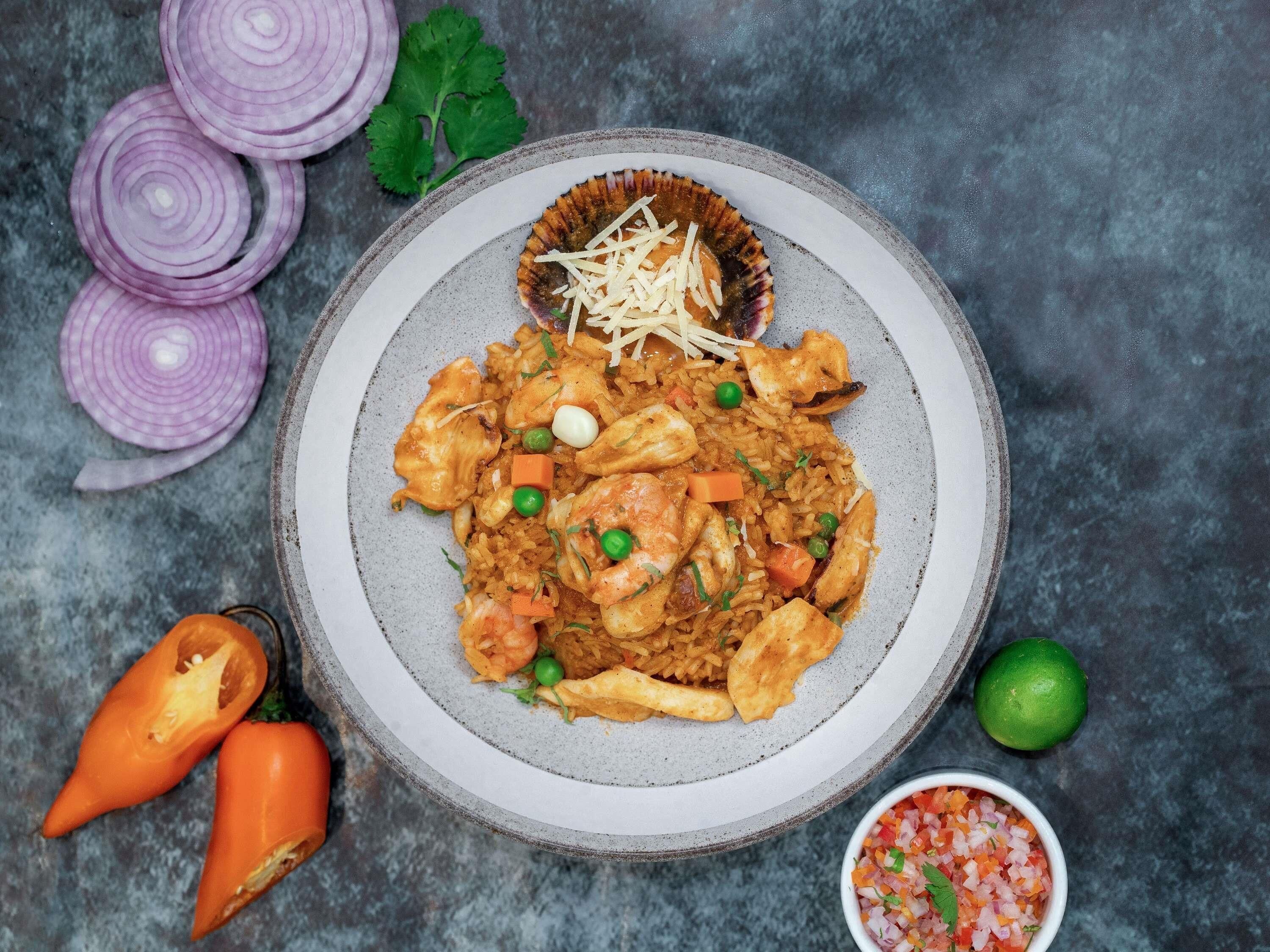 A plate of food with rice, vegetables, and meat, surrounded by sliced onions, a lime, and a bowl of chopped vegetables.