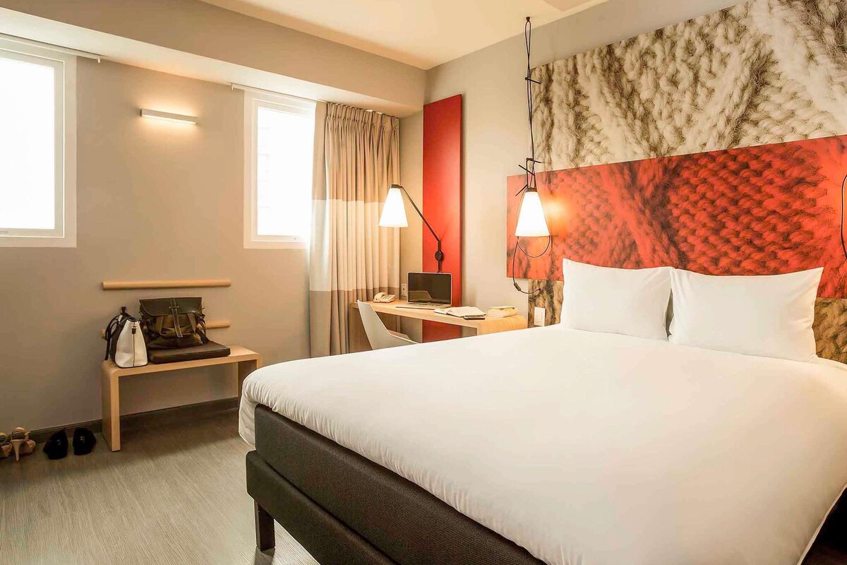 A hotel room with a large bed, a desk with a laptop, a chair, a lamp, and a window with curtains.