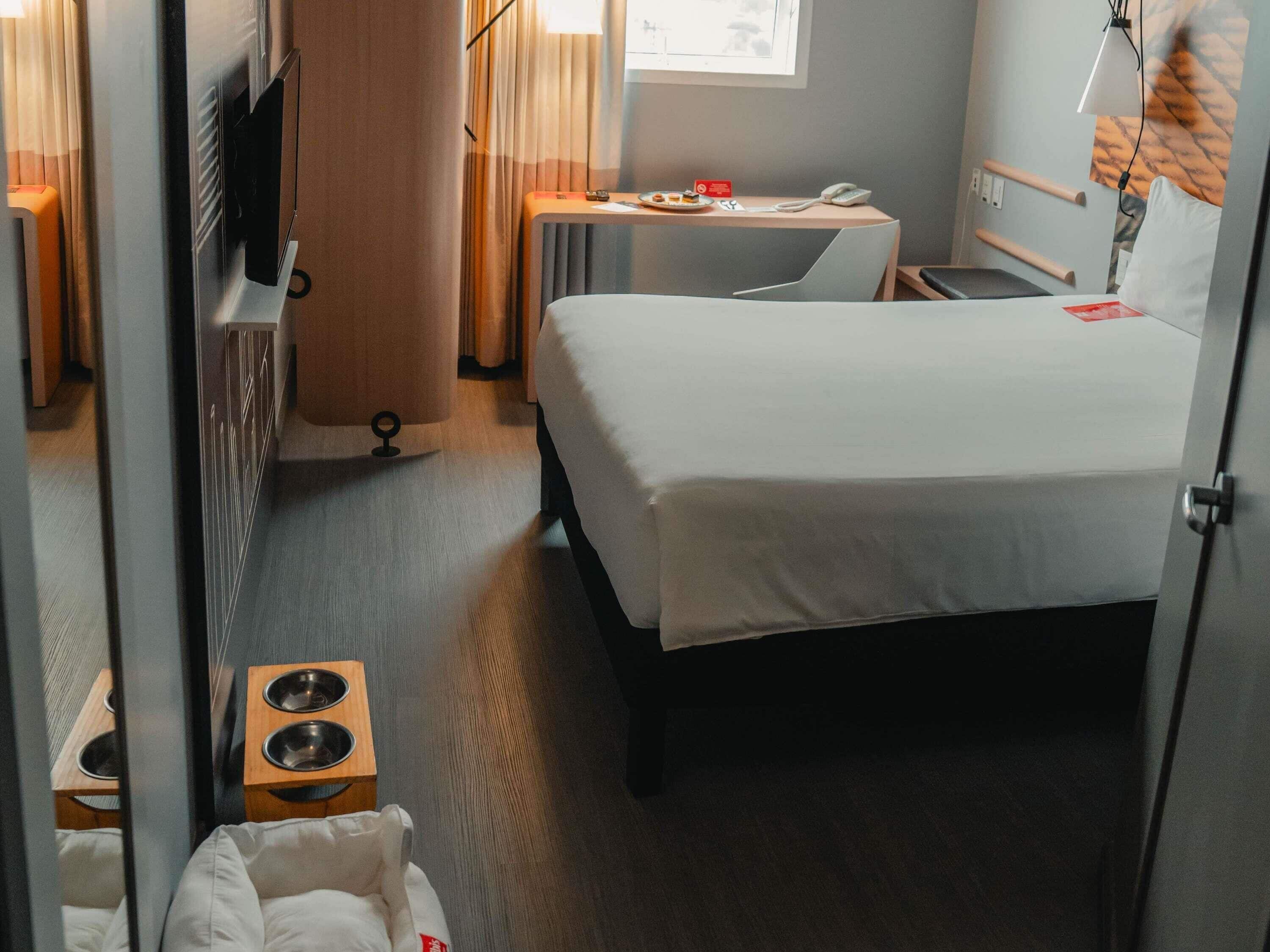 Habitación de hotel con cama, escritorio, televisión, ventana con cortinas, lámpara de pared y una mesita con un plato y un teléfono.