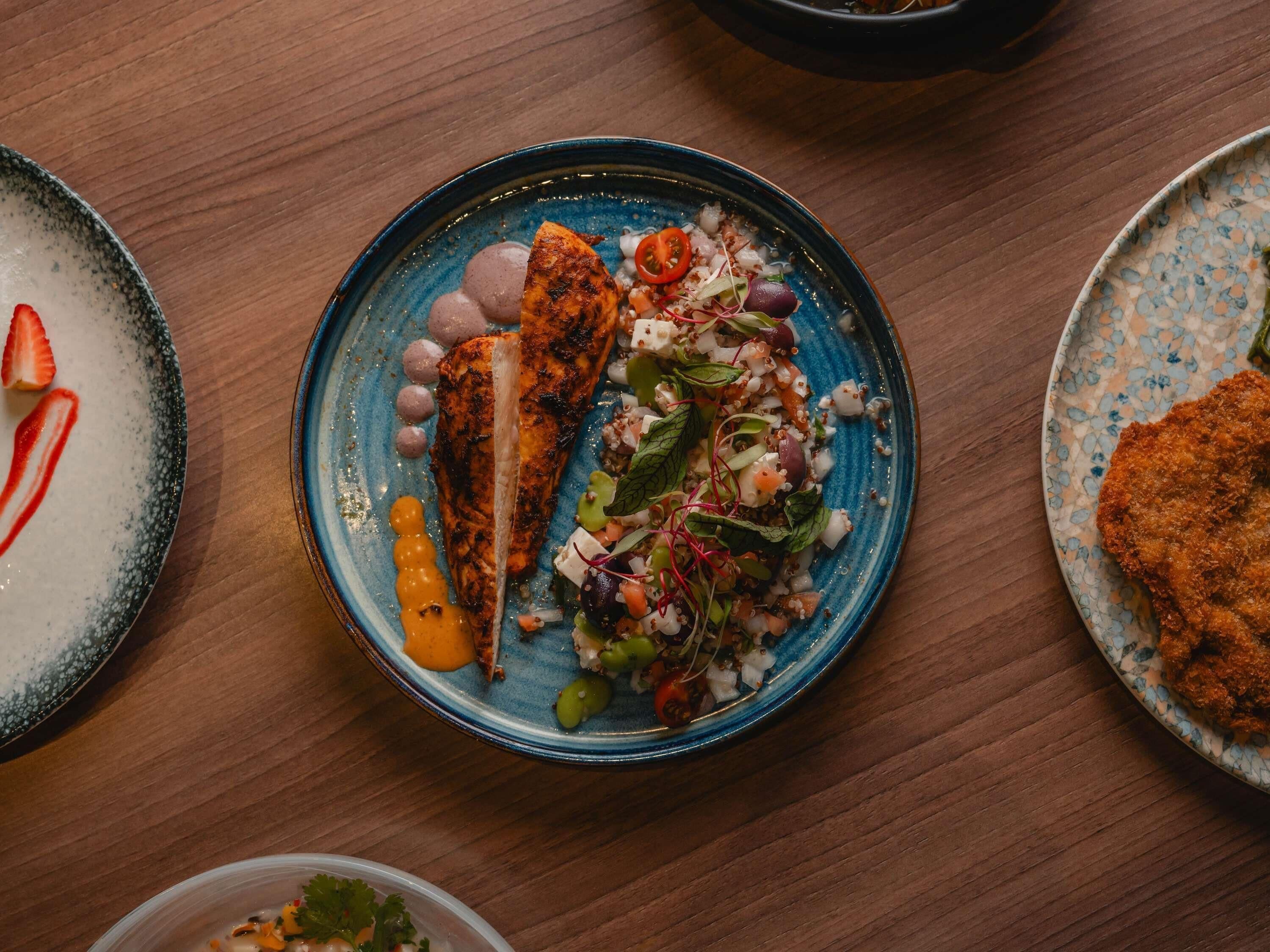 Una mesa de comedor con diversos platos, incluyendo un plato azul con pescado a la parrilla y una ensalada, un plato con algo empanizado y un plato con salsa blanca y fresas.