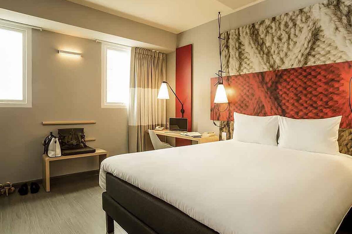 Habitación de hotel con una cama grande, un escritorio con una computadora portátil, una silla, una lámpara y una ventana con cortinas.
