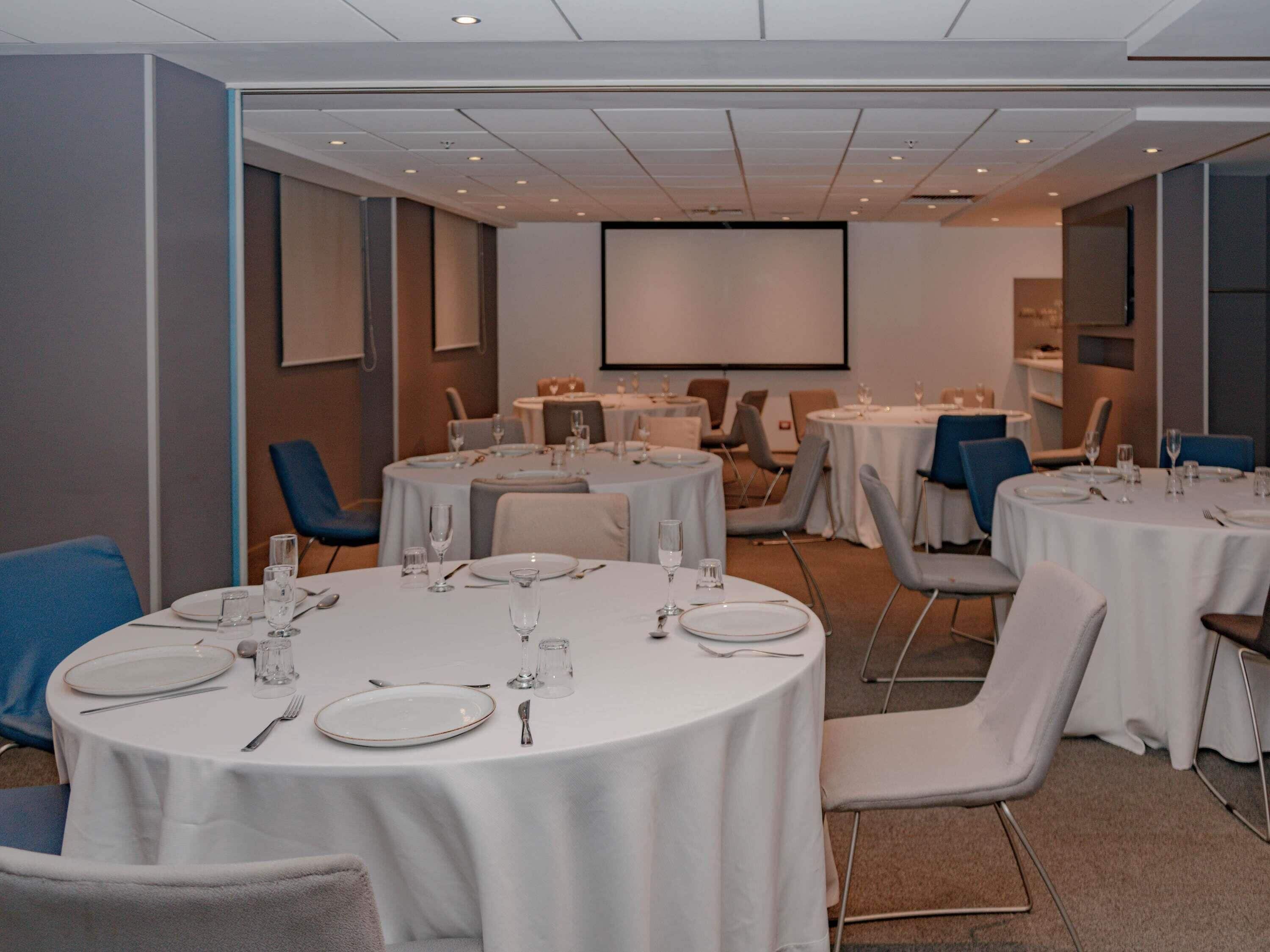 Un salón de conferencias con mesas redondas, sillas y una pantalla para proyector.
