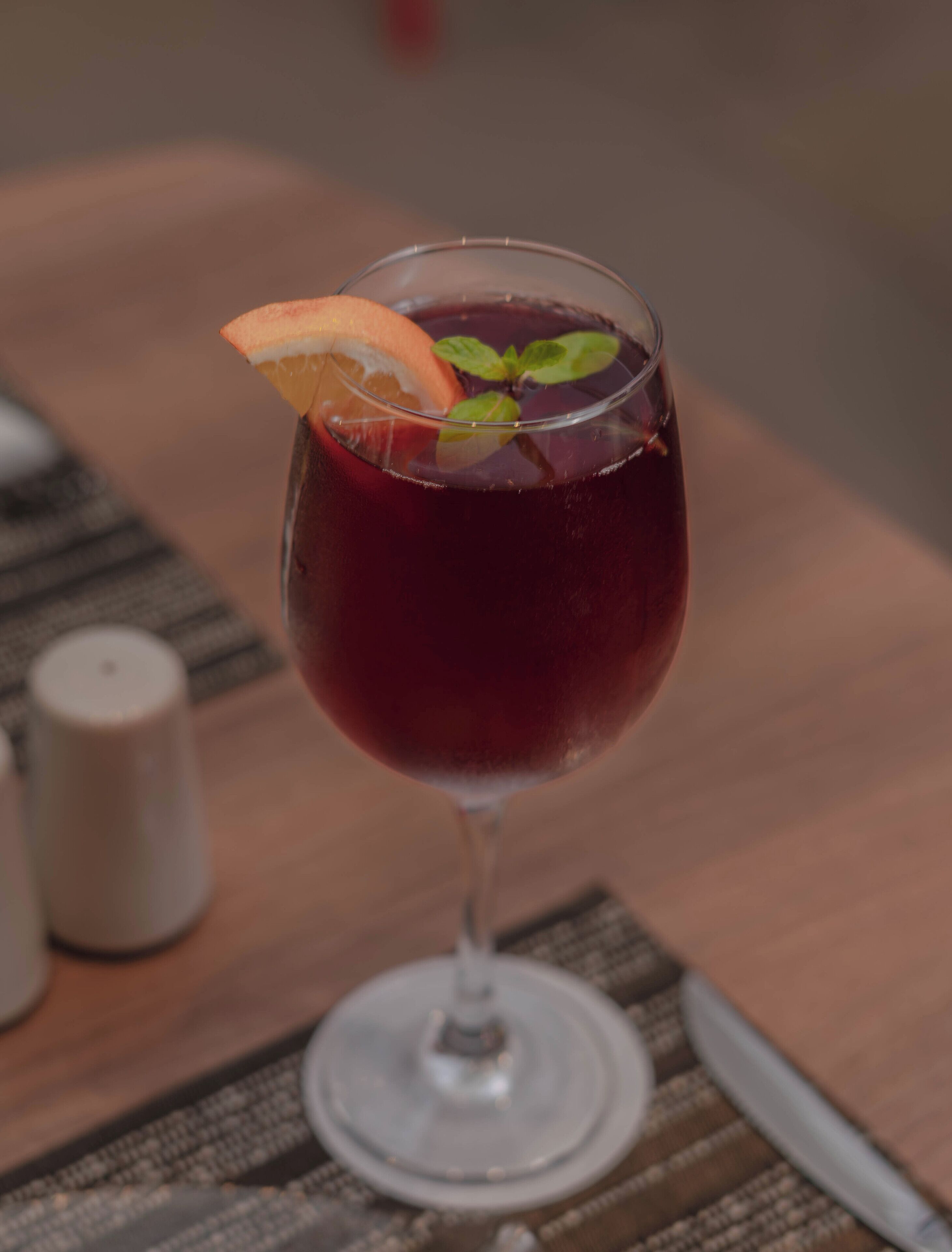 Un verre d’une boisson rouge, garni d’une tranche d’orange et de feuilles de menthe, posé sur une table en bois.