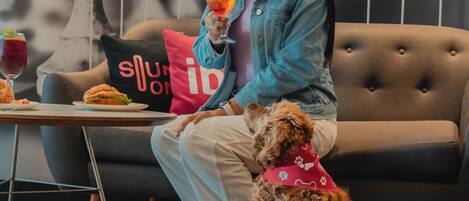 A person sitting on a sofa with a dog, holding a drink, and a table with food and drinks in front of them.