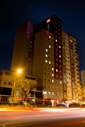 Un hotel de varios pisos con señalización iluminada, una calle en primer plano con estelas de luz y un cielo nocturno despejado.