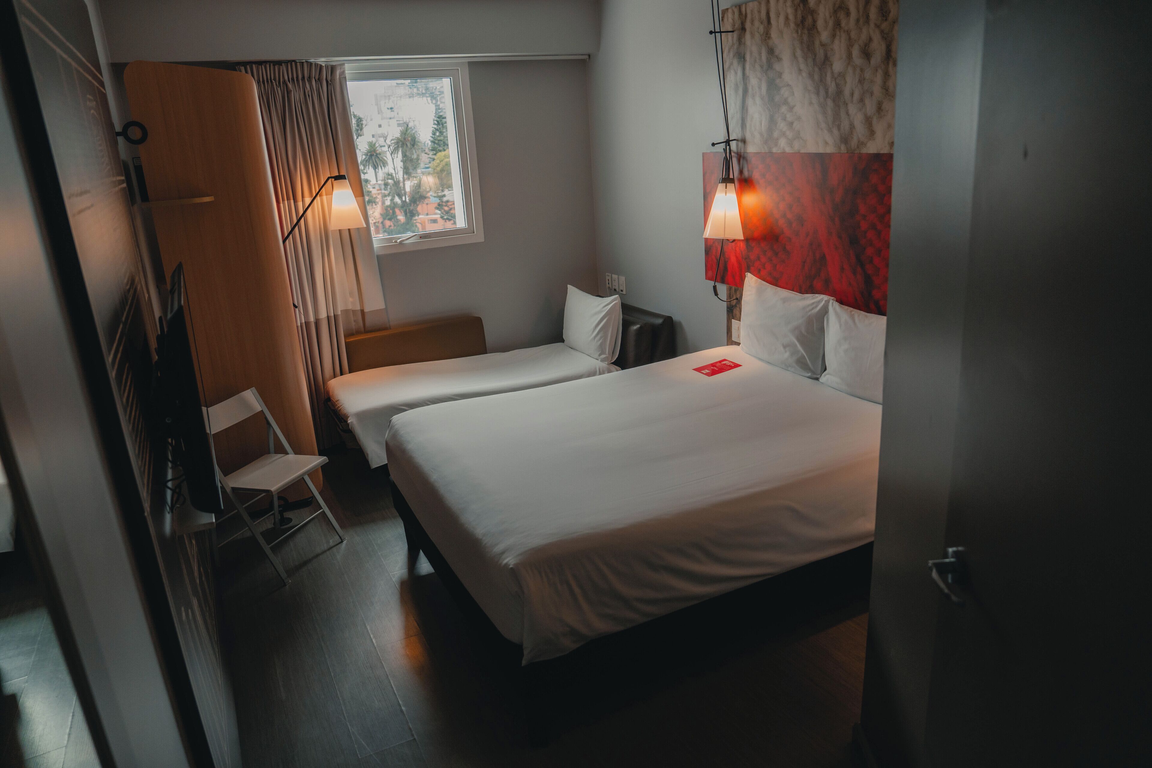 A hotel room with two beds, a chair, a window with curtains, and a lamp.