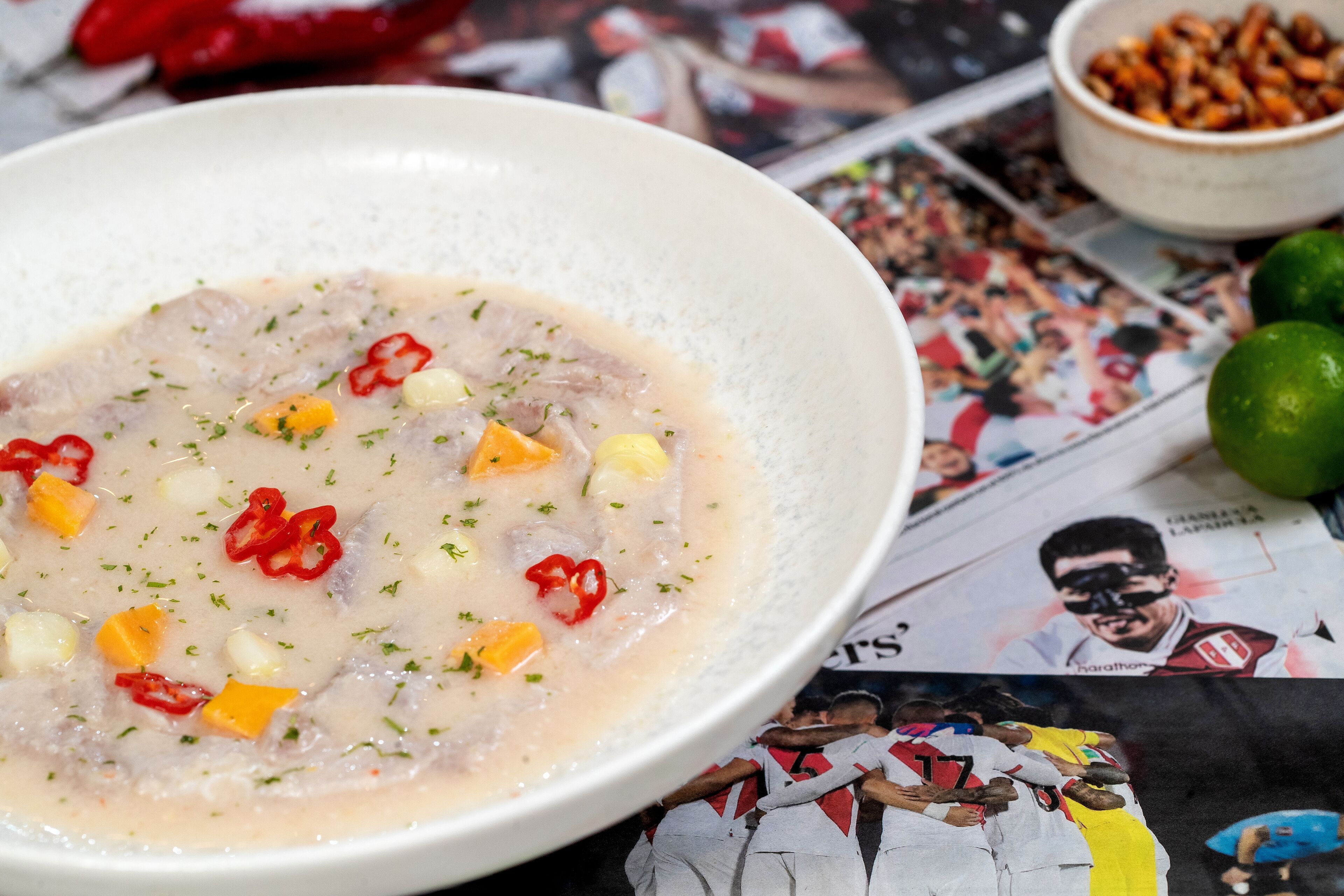 Un tazón de sopa con ingredientes visibles como maíz, pimientos rojos y hierbas, colocado sobre una mesa con periódicos y un cuenco de nueces.