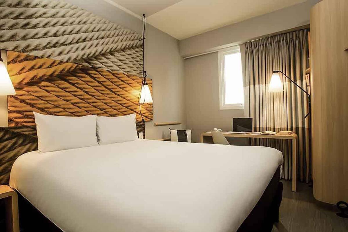 A hotel room with a large bed, a desk with a laptop, a chair, and a window with curtains.