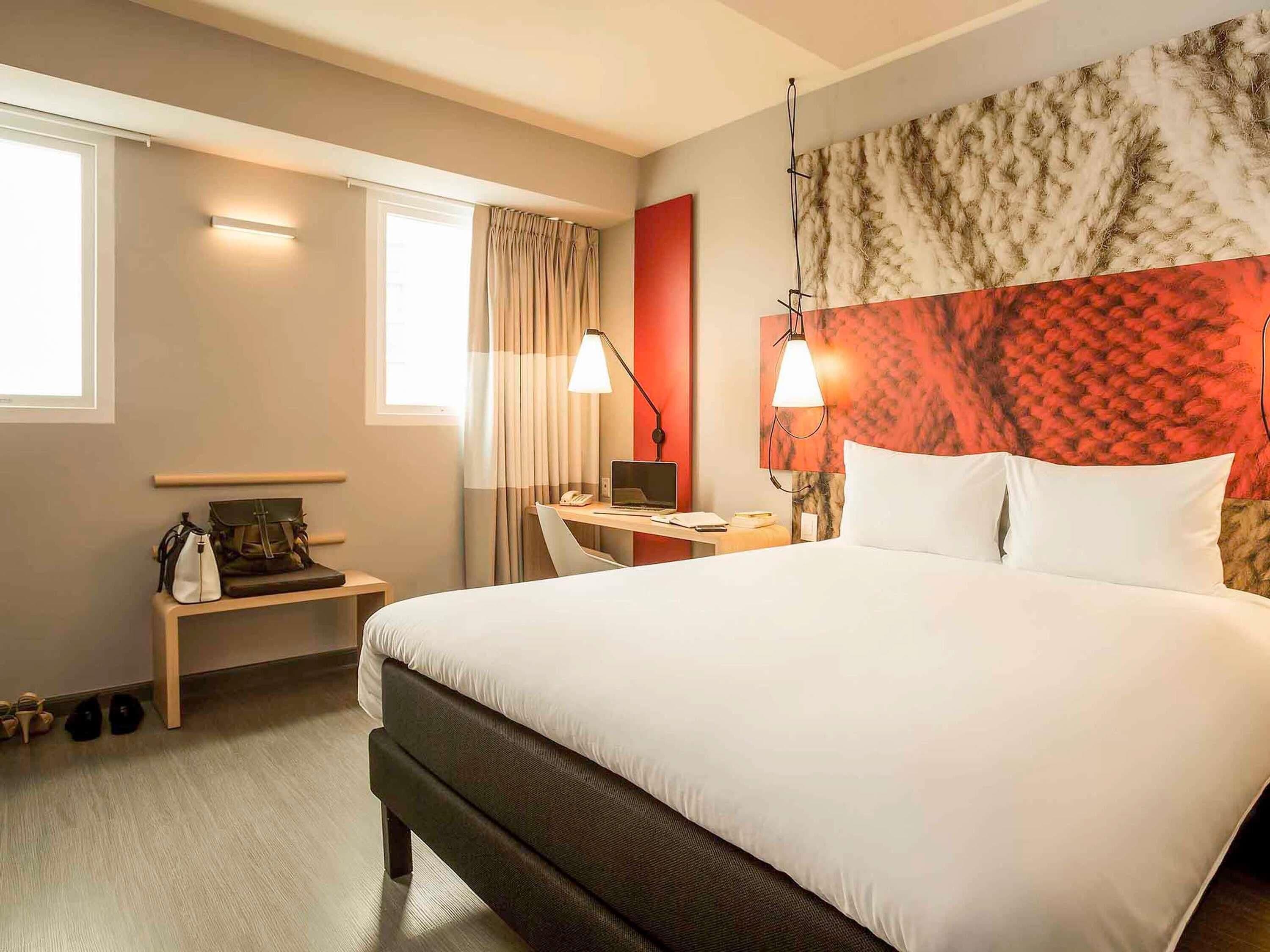Habitación de hotel con una cama grande, un escritorio con una computadora portátil, una silla, una lámpara y una ventana con cortinas.