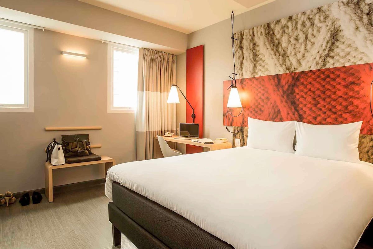 Ein Hotelzimmer mit einem großen Bett, einem Schreibtisch mit einem Laptop, einem Stuhl, einer Lampe und einem Fenster mit Vorhängen.
