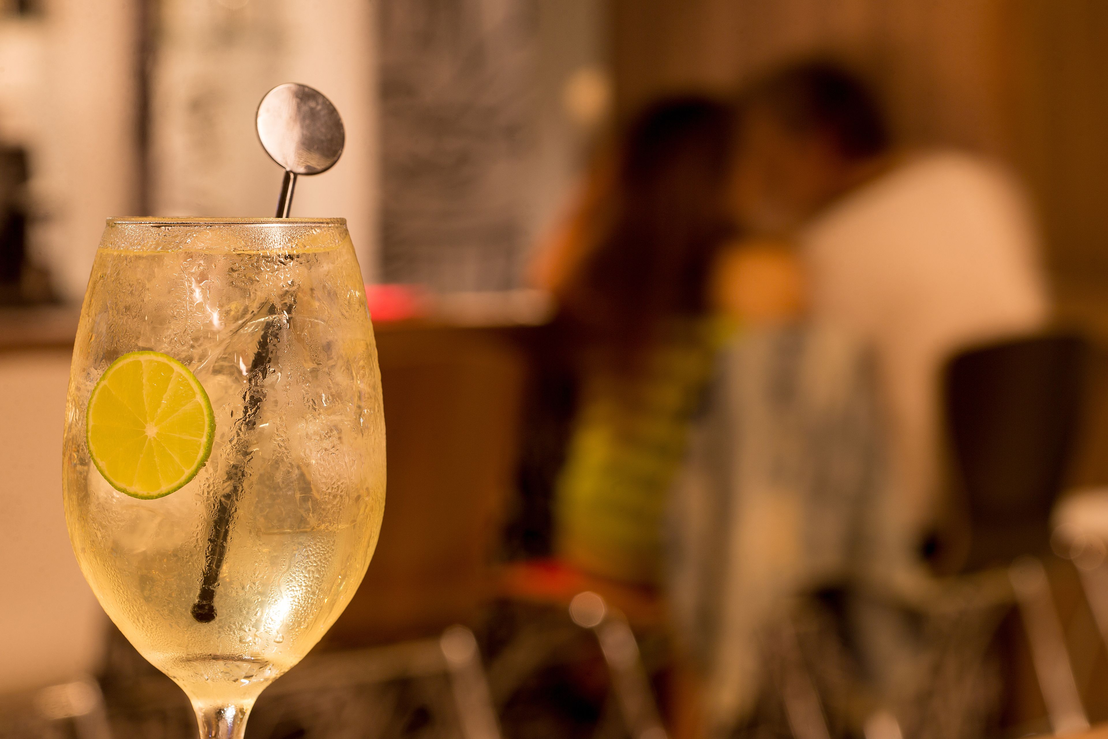 A glass of a clear beverage with a lime slice and a metal stirrer, placed on a bar counter with a blurred background of people.