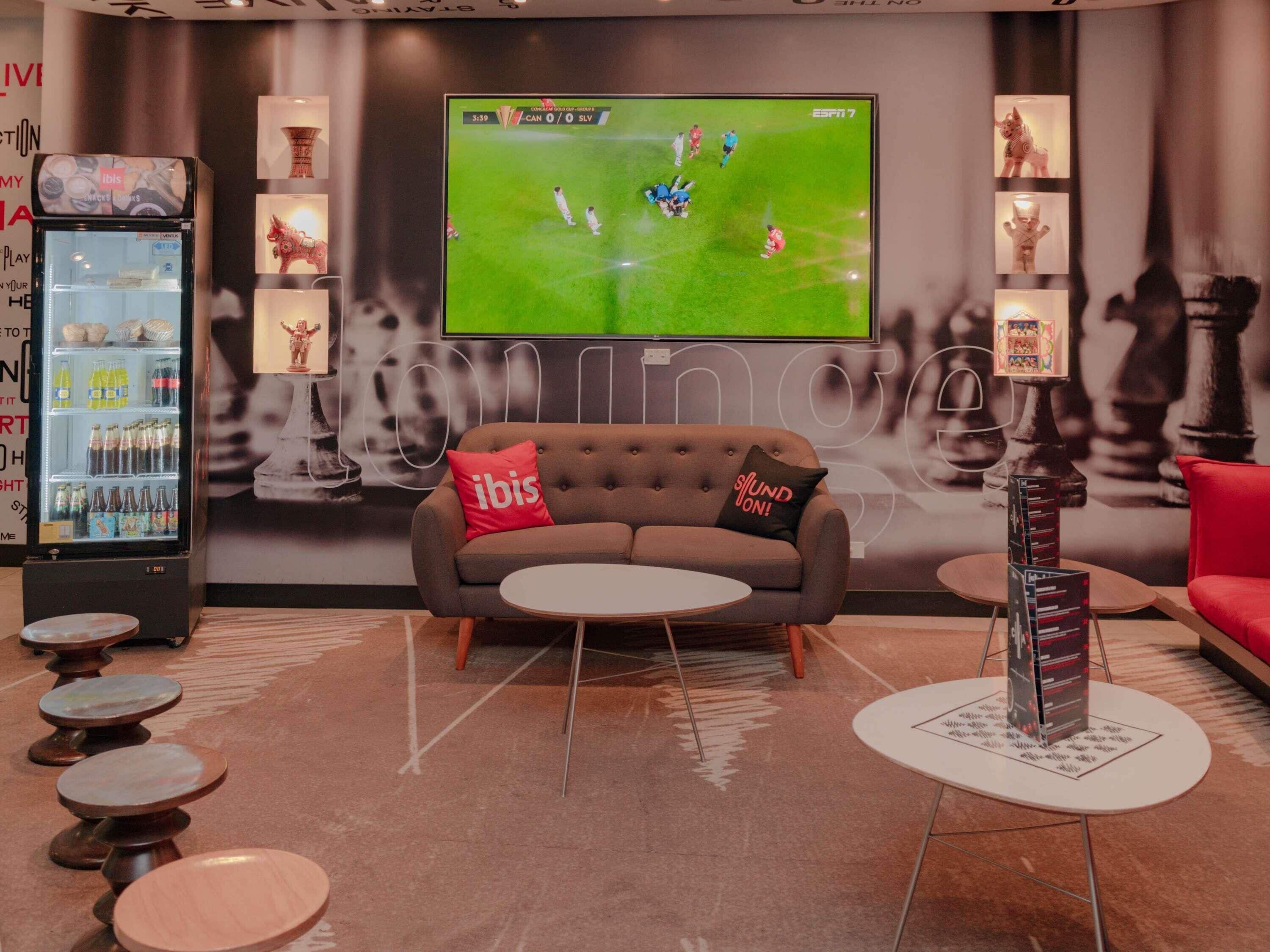 A modern lounge area with a sofa, a small round table, a beverage dispenser, and a television displaying a sports game.