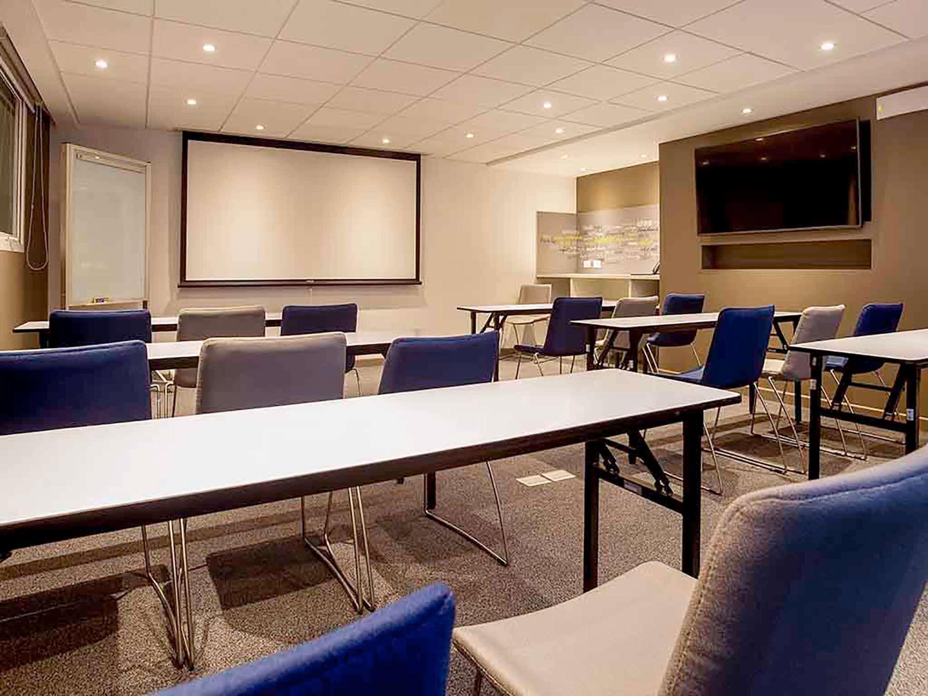 A conference room with tables, chairs, a projector screen, and a TV.