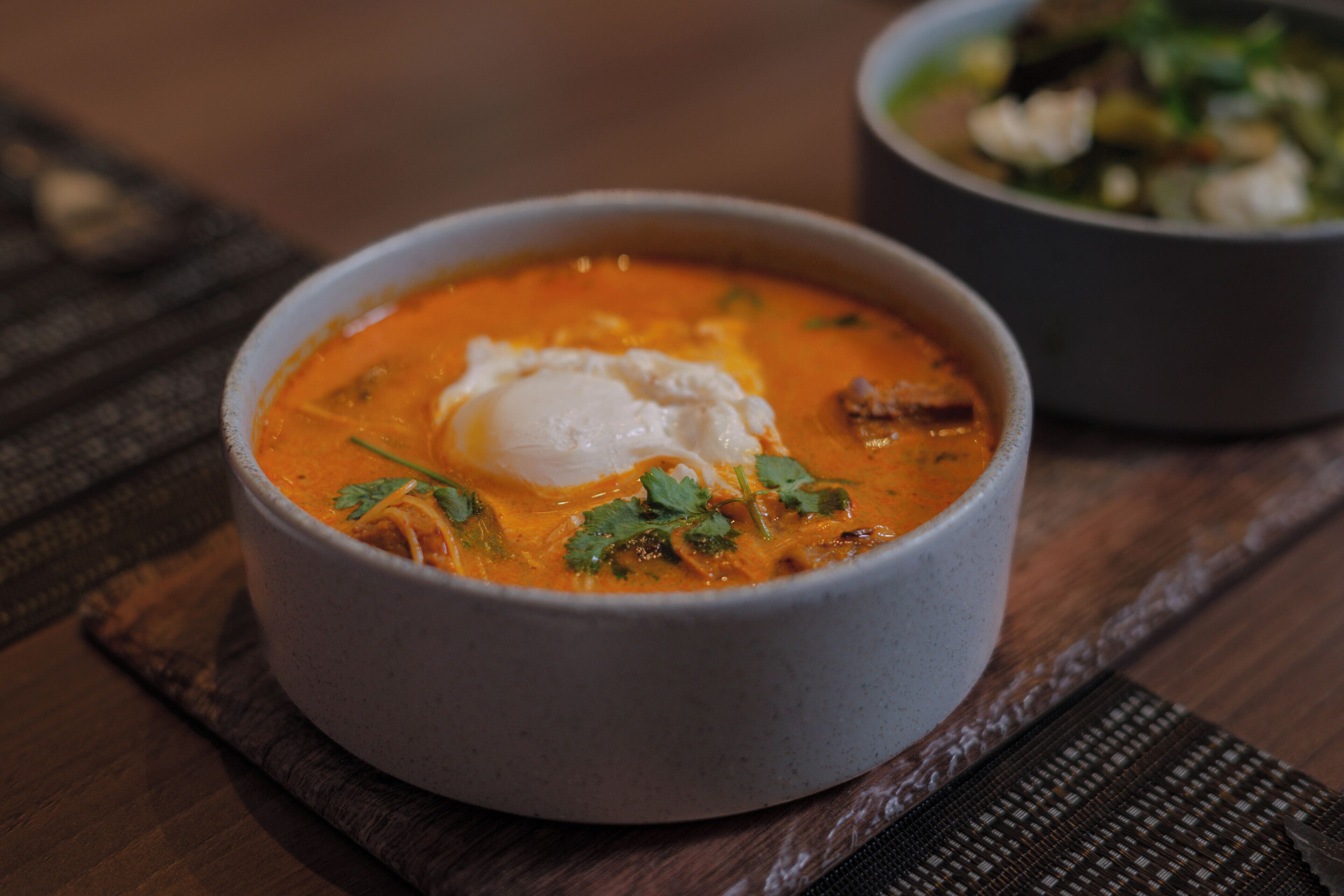 Un tazón de sopa con un huevo escalfado y cilantro como decoración, sobre una mesa de madera.