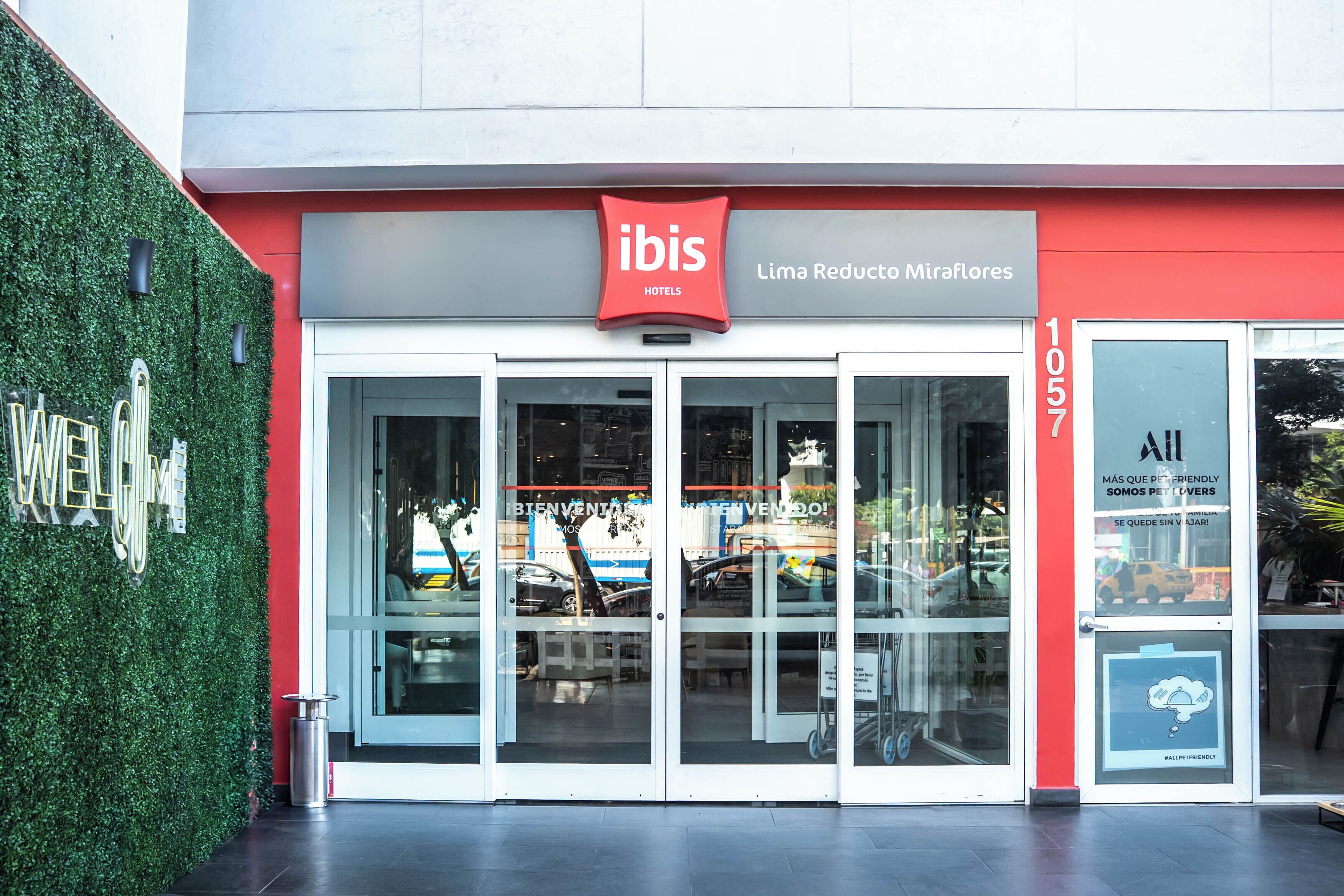 A hotel entrance with a red and white facade, a green wall with a 'WELCOME' sign, and a glass door with a sign reading 'ibis Hotels Lima Reducto Miraflores'.
