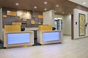 A reception desk with a digital display, a refrigerator, and a patterned wall. - Holiday Inn Express & Suites Brookshire - Katy Freeway by IHG (Brookshire)