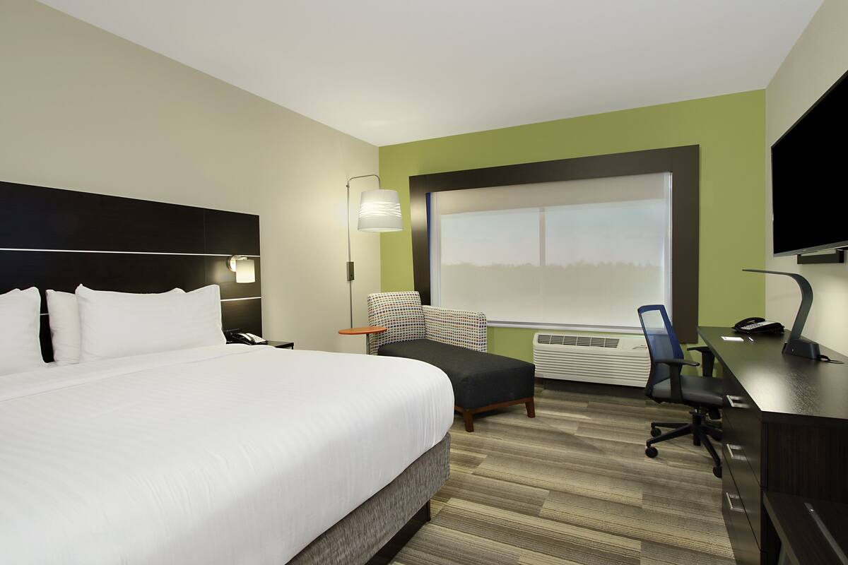 A hotel room with a large bed, a desk with a computer, a chair, a lamp, and a large window.