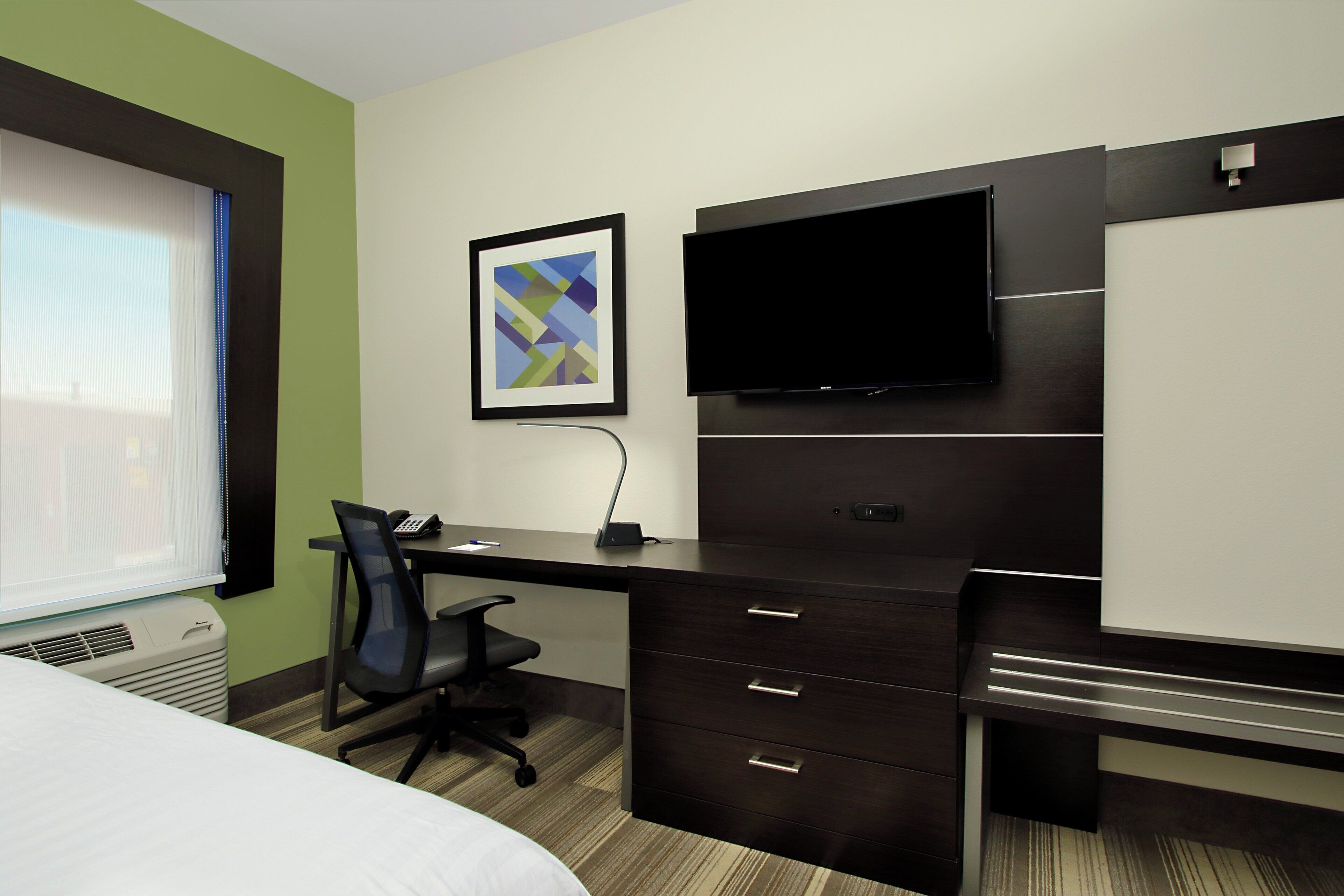 A hotel room with a bed, a desk, a chair, a flat-screen TV, a framed abstract art piece, and a window with blinds.