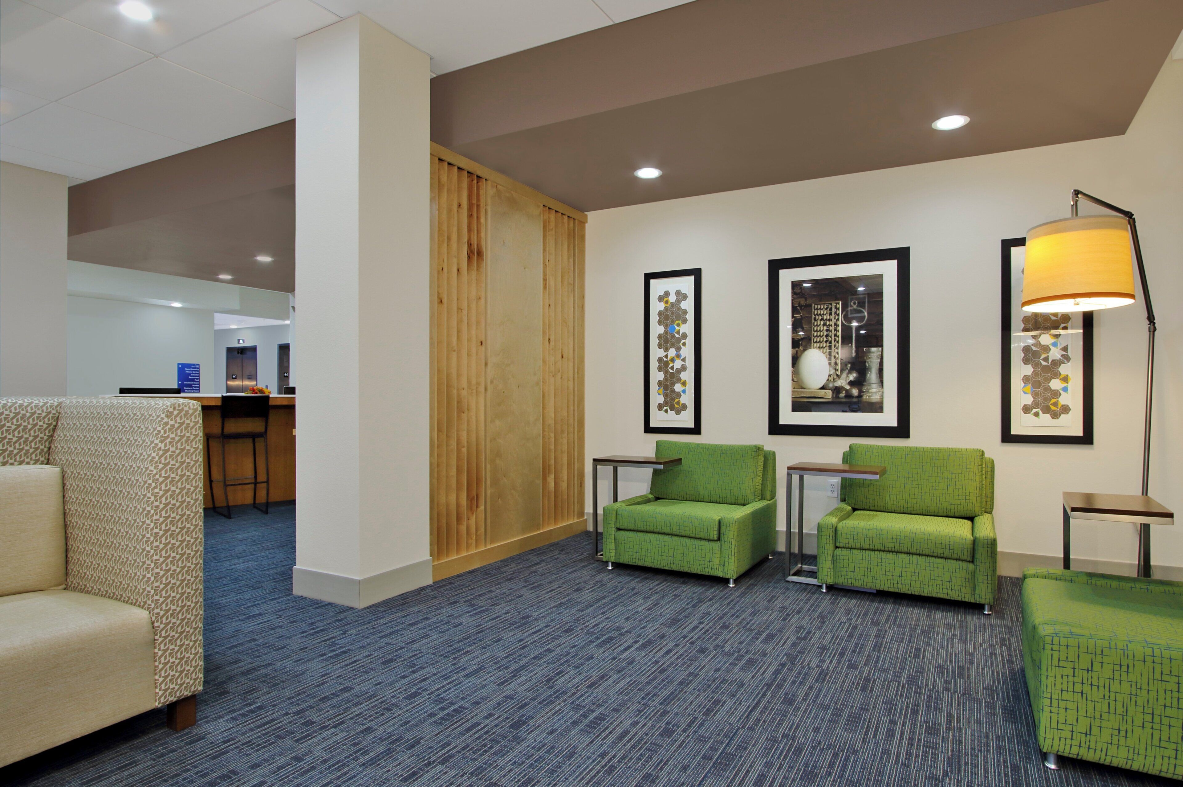 A hotel lobby with a reception desk, two green armchairs, a patterned sofa, and framed artwork on the walls.