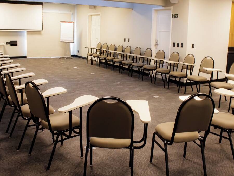 Un salón de conferencias con filas de sillas y mesas, una pantalla de proyector y una puerta.