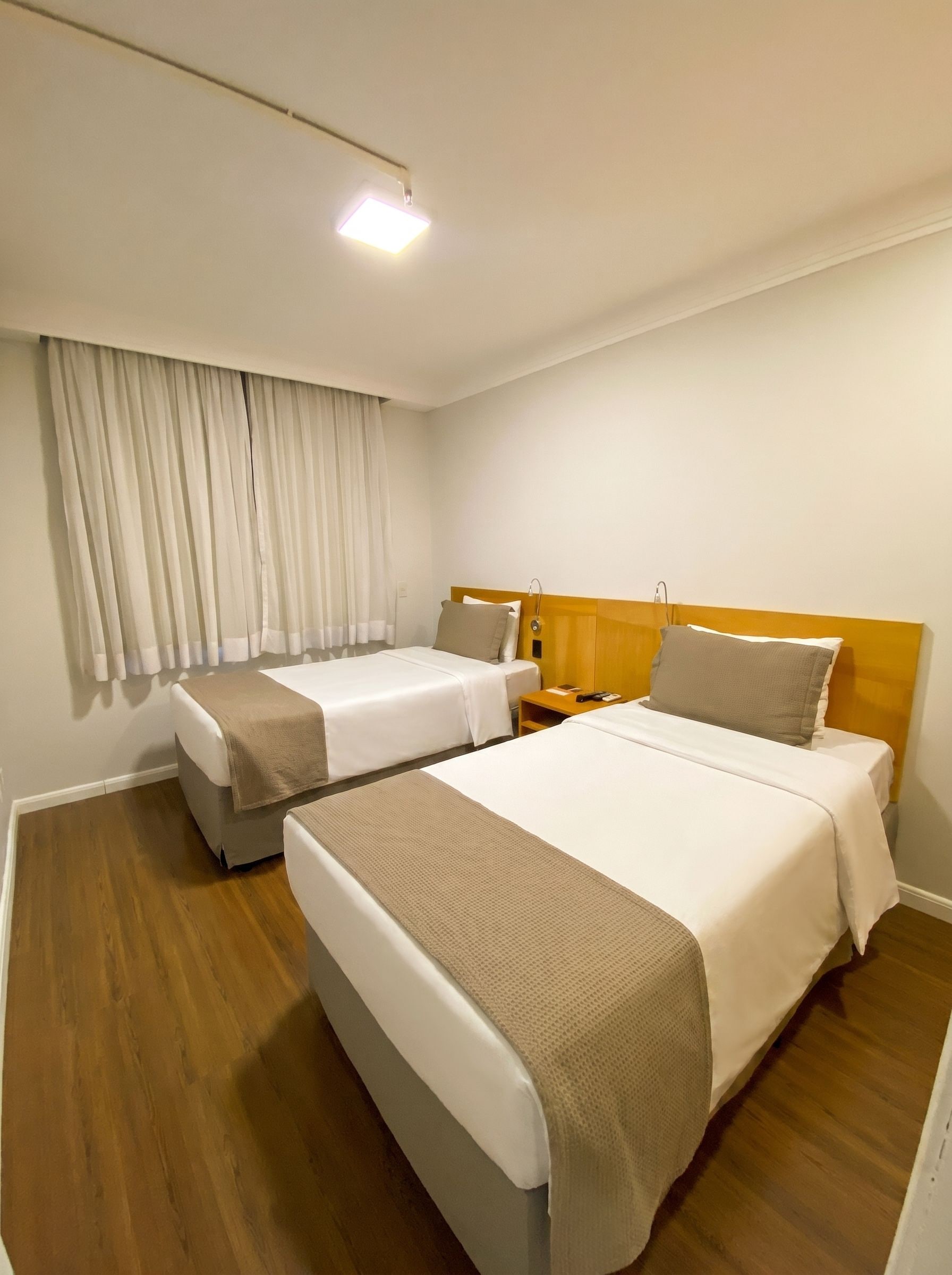 Ein Hotelzimmer mit zwei Betten, einem Holz-Kopfteil, einem Nachttisch, einer Lampe und einem fenster mit Vorhängen.
