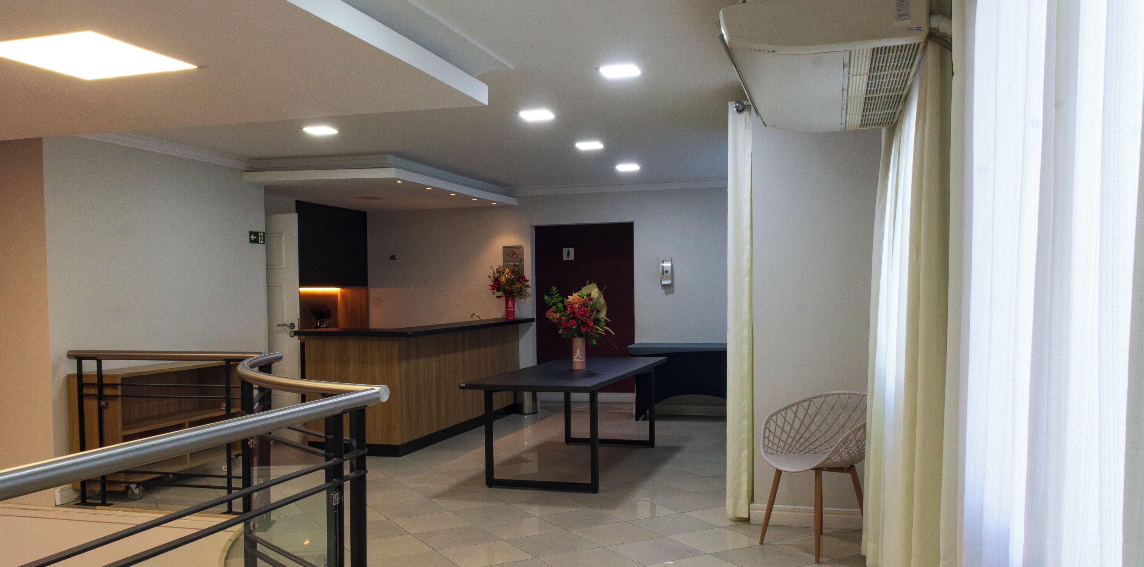 A hotel lobby with a reception desk, a seating area with a table and chairs, a staircase with a handrail, and a wall-mounted air conditioning unit.