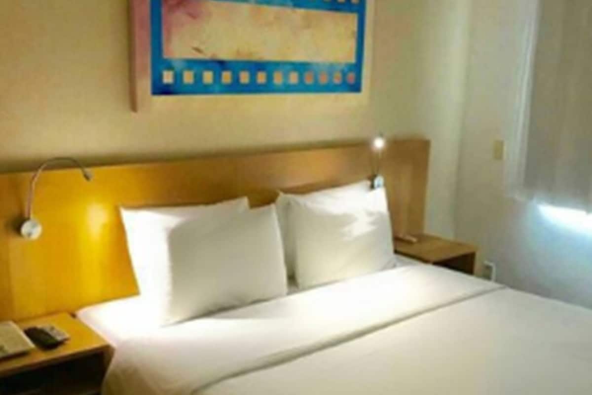 A hotel room with a bed, a nightstand with a lamp, a bedside table with a phone, and a wall-mounted painting.