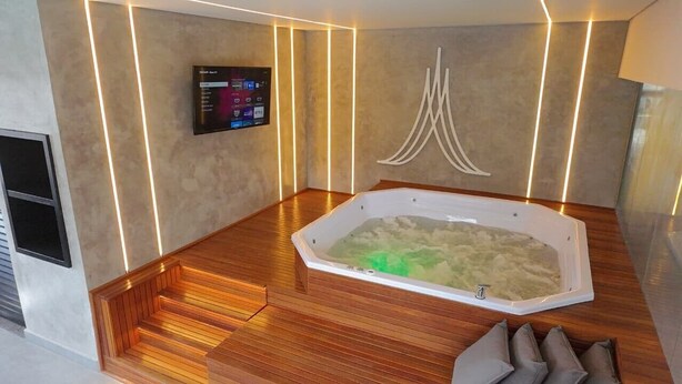 A modern bathroom with a freestanding bathtub, wooden flooring, a wall-mounted TV, and a unique wall art piece. - Marsala Apart Hotel (Mogi das Cruzes)