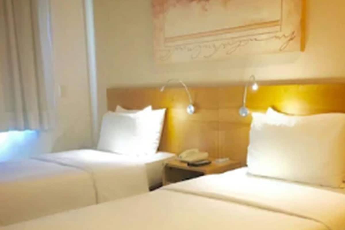 A hotel room with two beds, a nightstand with a phone, and a wall-mounted lamp.